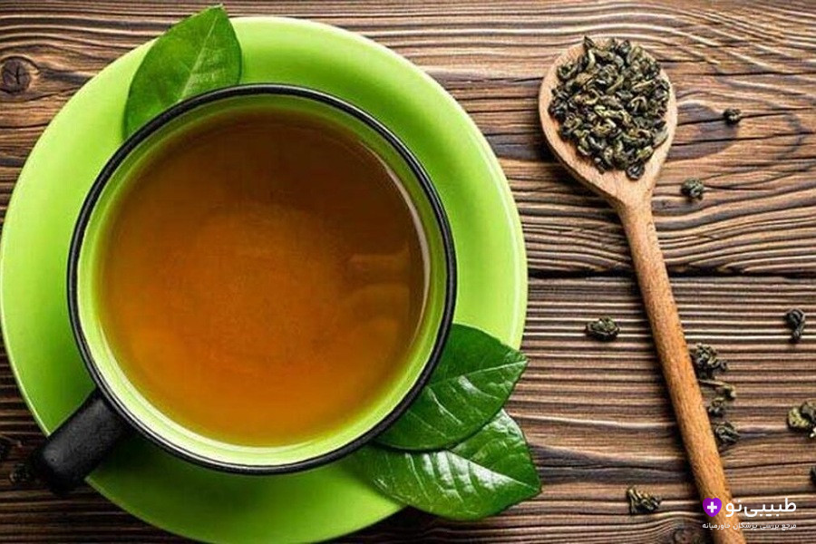 فنجان بخارآلود چای سبز در نعلبکی سبز پررنگ، با تزیین برگ‌های سبز تازه و دانه‌های چای شل روی قاشق چوبی، قرارگرفته روی میز چوبی روستایی