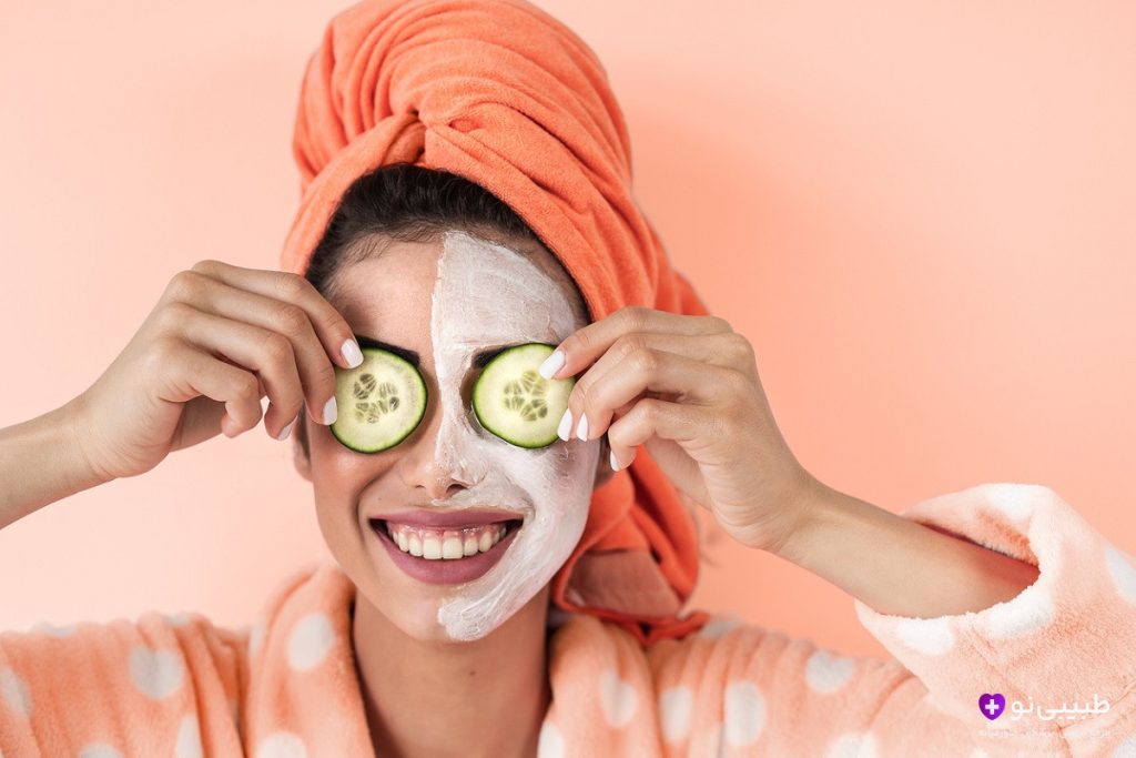 دختر جوان شاد با حوله نارنجی دور سر و ردای خال‌خالی، ماسک صورت جواسناز پوست زده و دو برش خیار روی چشم‌هاش گذاشته، با لبخند بزرگ به دوربین نگاه می‌کنه.
