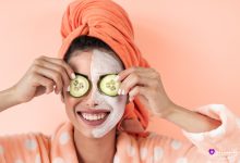 دختر جوان شاد با حوله نارنجی دور سر و ردای خال‌خالی، ماسک صورت جواسناز پوست زده و دو برش خیار روی چشم‌هاش گذاشته، با لبخند بزرگ به دوربین نگاه می‌کنه.