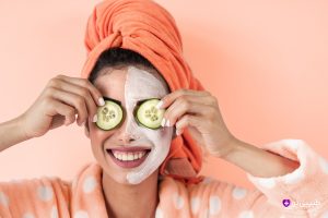 دختر جوان شاد با حوله نارنجی دور سر و ردای خال‌خالی، ماسک صورت جواسناز پوست زده و دو برش خیار روی چشم‌هاش گذاشته، با لبخند بزرگ به دوربین نگاه می‌کنه.
