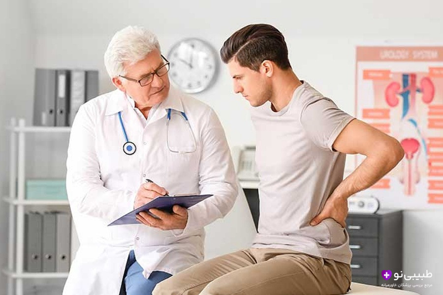 پزشک مسن با روپوش سفید در حال یادداشت‌برداری در حالی که بیمار جوان دستش را روی کمر گذاشته است.