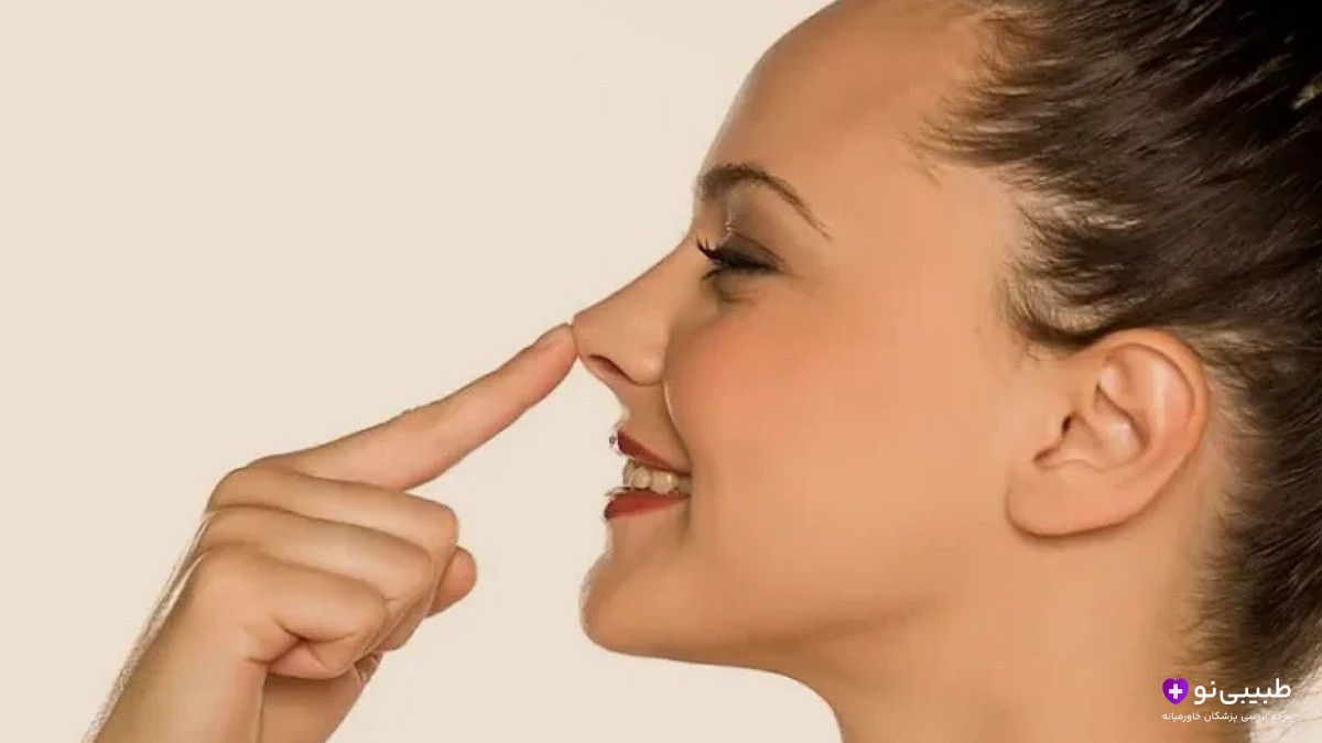 زن جوان با لبخند ملایم، در حال لمس نوک بینی خود با انگشت اشاره، نیمرخ از کنار، پسزمینه سفید و روشن.