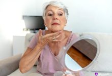 خانم مسن در حال بررسی ناحیه گردن و فک خود با آینه دستی برای بررسی نشانه‌ای از سفتی بیش‌ازحد یا عدم تقارن احتمالی پس از لیفت صورت و گردن.