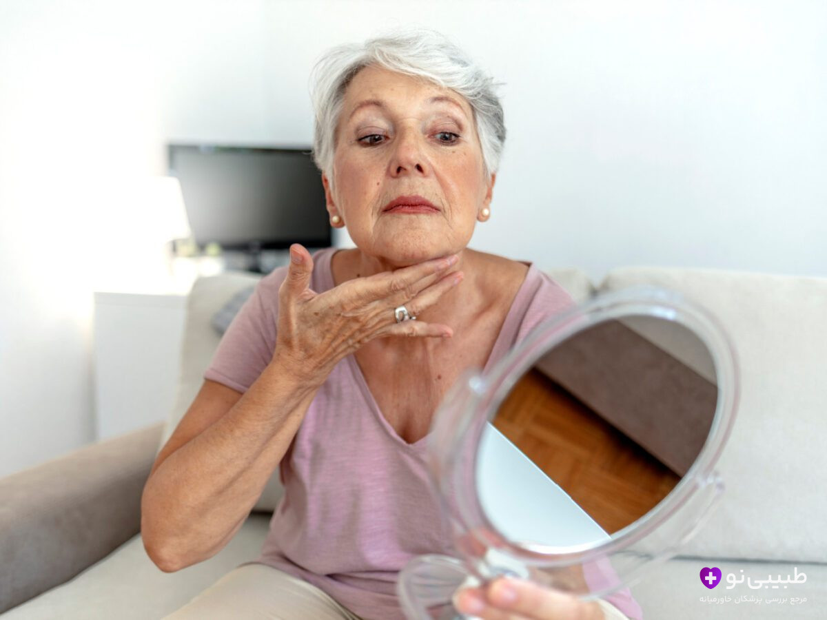 خانم مسن در حال بررسی ناحیه گردن و فک خود با آینه دستی برای بررسی نشانهای از سفتی بیشازحد یا عدم تقارن احتمالی پس از لیفت صورت و گردن.